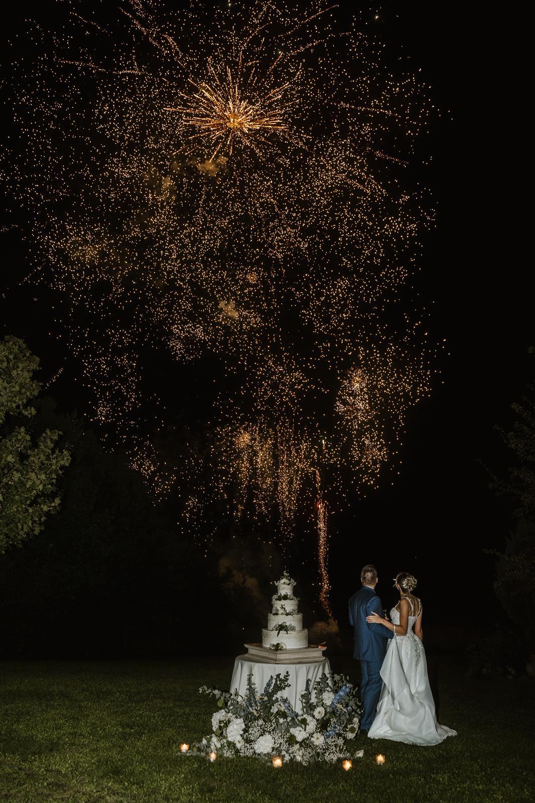 Coppia di sposi che osserva il cielo pieno di fuochi d'artificio con la torta nuziale in primo piano.